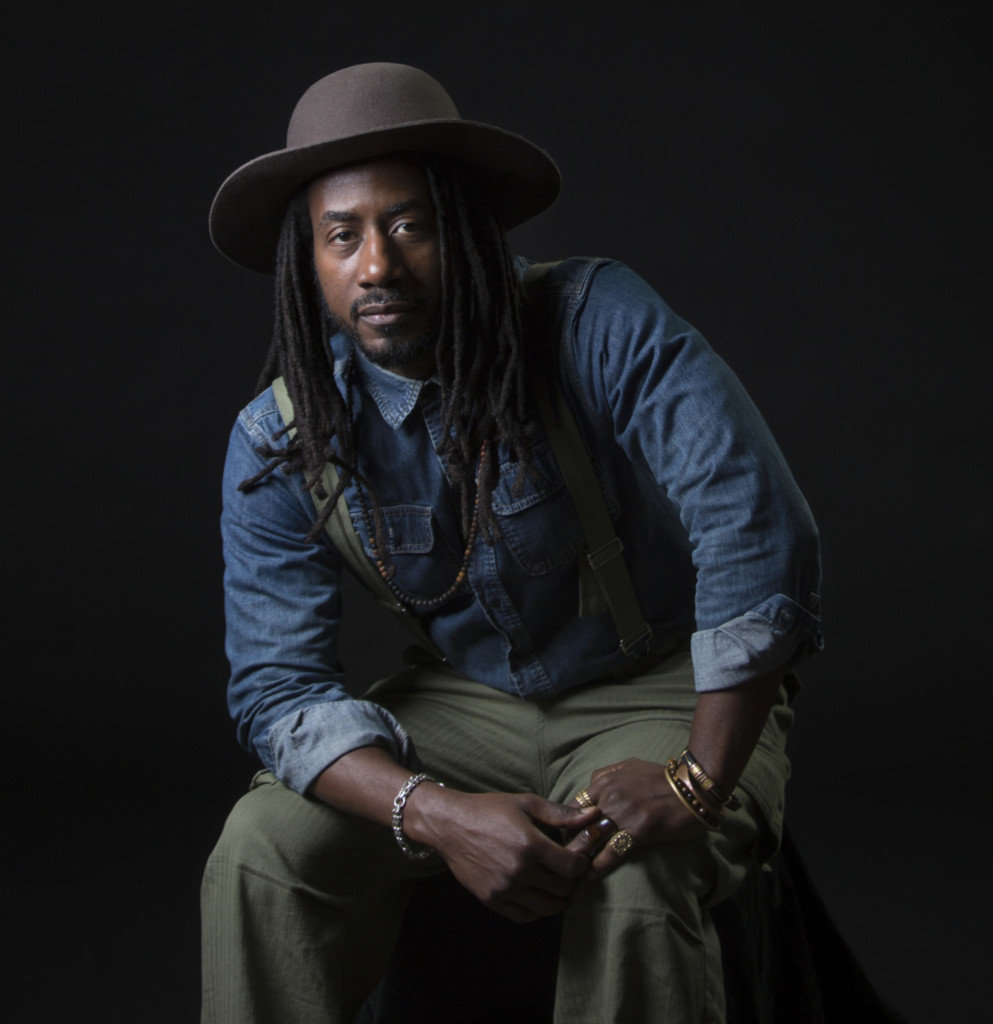 A Black man is sitting against a black backdrop. He is wearing a hat, denim shirt, green overalls, and jewelry.