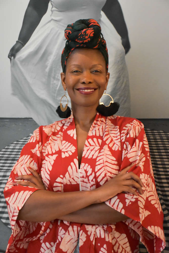 A Black woman wearing a red and white patterned top, a headwrap, and big earrings smilingat the camera. Her arms are crossed and she is standing in front of a black and white image.