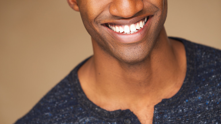 A Black man is smiling straight at the camera. He is wearing a dark blue button up shirt and is against a brown wall.