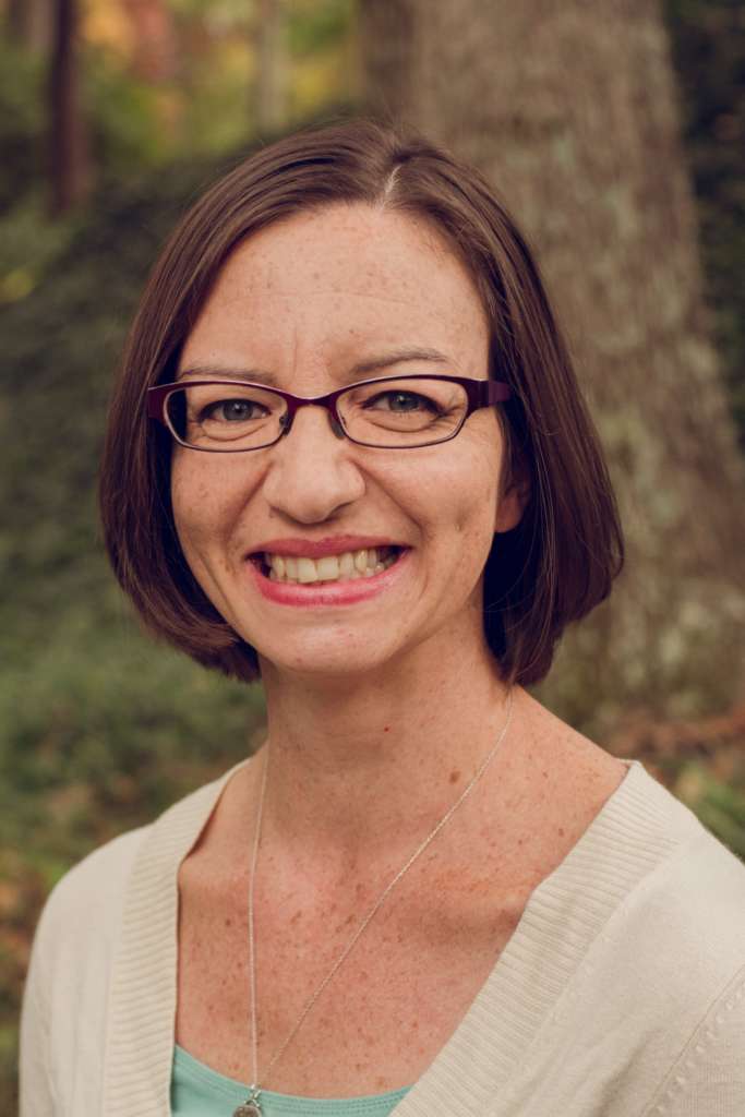 A white woman with glasses and dark hair is smiling at the camera. She is wearing a blue top with a white sweater and a long necklace. She is standing against a backdrop of trees.