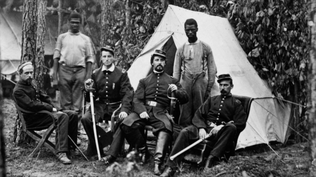 A black and white photo from the Civil War of four white male soldiers sitting down. Behind them are two Black men standing. They are outside with a tent in the background.