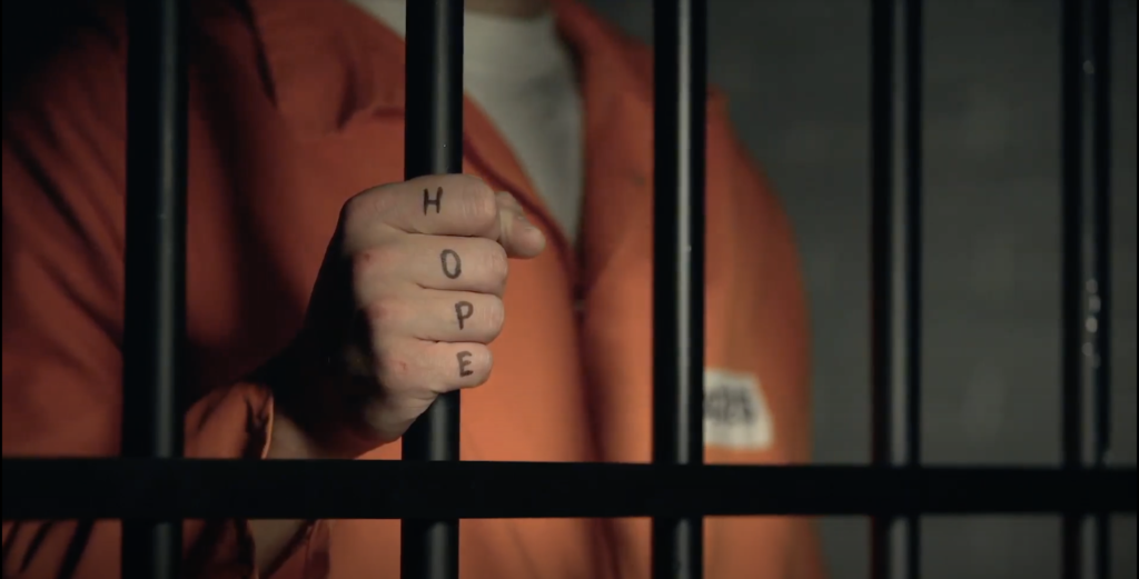 A person in an orange prison jumpsuit holds the cell bars and their fist has "hope" written on it