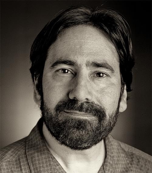 Joel Silverman is smiling at the camera in a black-and-white photo, wearing a plaid button up shirt.