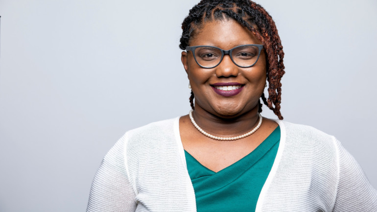 Amina S. McIntyre is smiling in front of a white backdrop wearing a teal shirt, white cardigan, glasses, and pearl necklace.