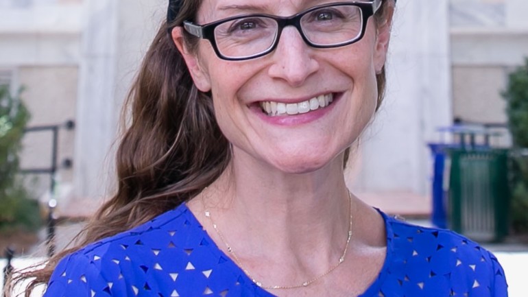 Ellie Schainker is smiling in front of a building outdoors. She is wearing glasses, a blue top, and a gray headband