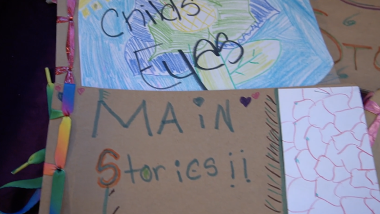 Cardboard books with writing in marker saying "main stories" and a sheet of paper saying "childs eyes" on top of a crayon coloring of a flower